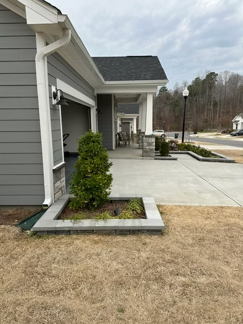 Gray house with porch, driveway, and small garden bed with a small tree. Overcast sky.