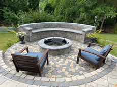 Round patio with built-in stone bench, fire pit, two wooden chairs; green grass, trees in background.