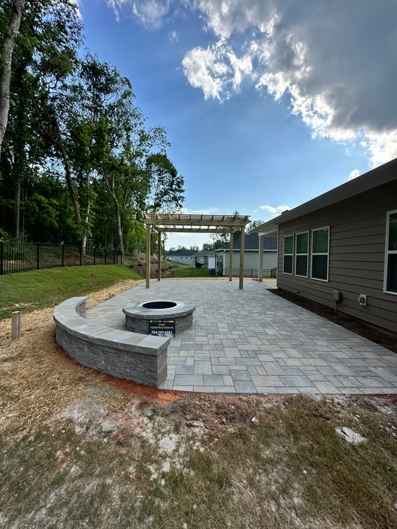 A patio with pavers, fire pit, and pergola next to a house under a cloudy sky.