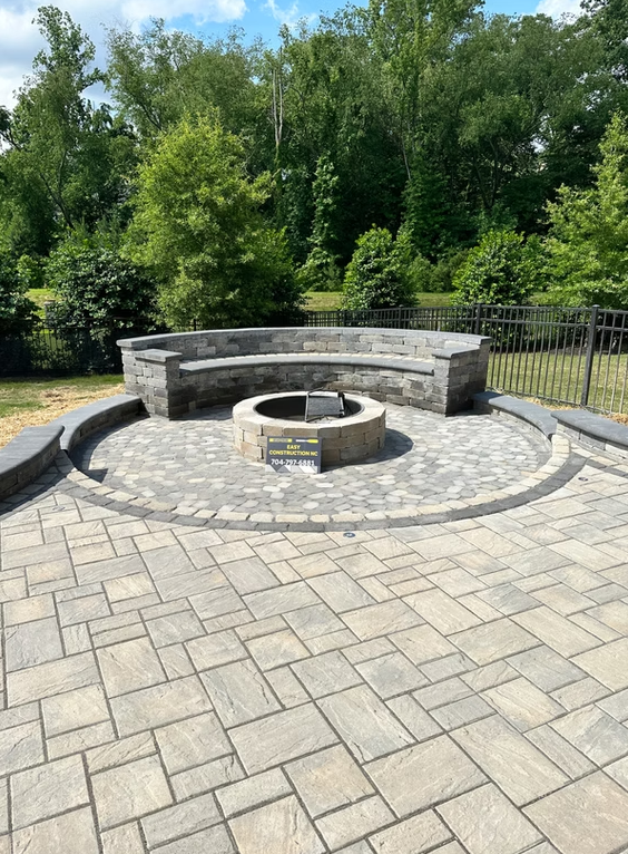 Circular stone patio with fire pit and built-in curved seating, surrounded by trees and a black fence.