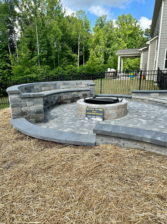 Stone patio with built-in seating surrounding a fire pit, set in a yard with trees and house.
