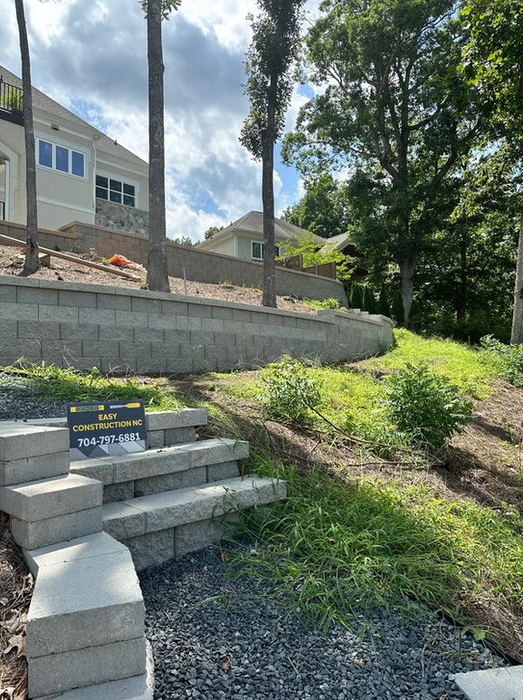 Steps and retaining wall in front of a light-colored house, with trees and greenery.