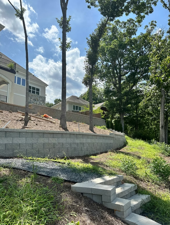 Stone retaining wall with steps, trees, and house under a blue sky with clouds.