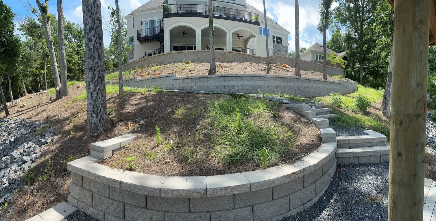 Retaining walls with steps lead to a large house on a hillside, trees surround the house.