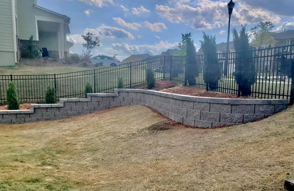 Stone retaining wall with steps, black fence, and evergreens in a yard under a partly cloudy sky.
