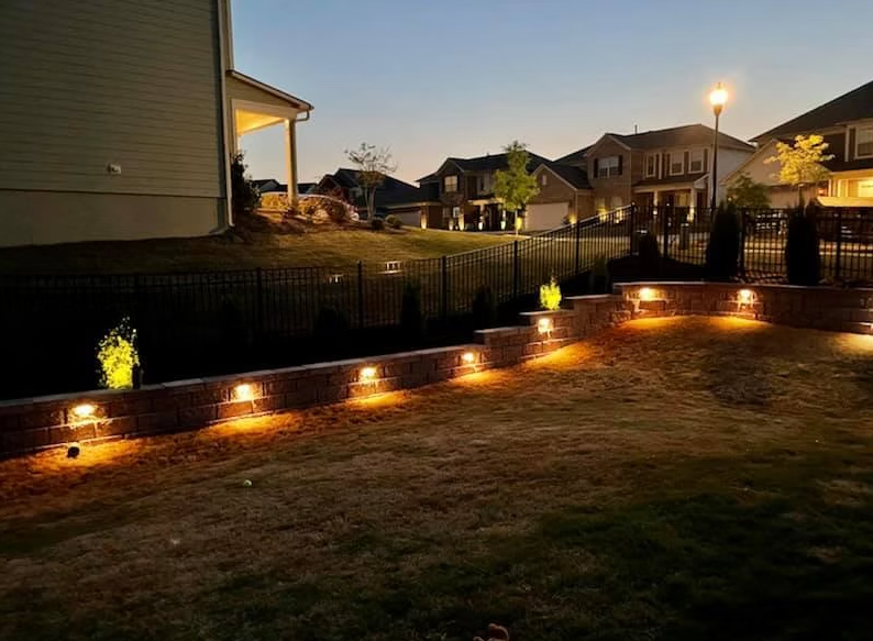Backyard at dusk with brick retaining walls, lit steps, and soft yellow lights. Houses in the background.