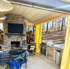 Outdoor stone fireplace with TV, grill, yellow curtains, and wooded view.