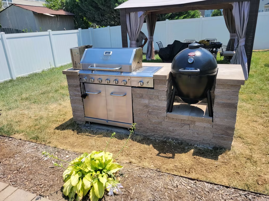 Outdoor grill station with stainless steel gas grill and black egg smoker under a canopy.