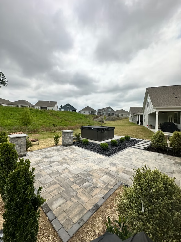 Patio with pavers, dark hot tub, black rock landscaping, green grass, and houses under a cloudy sky.