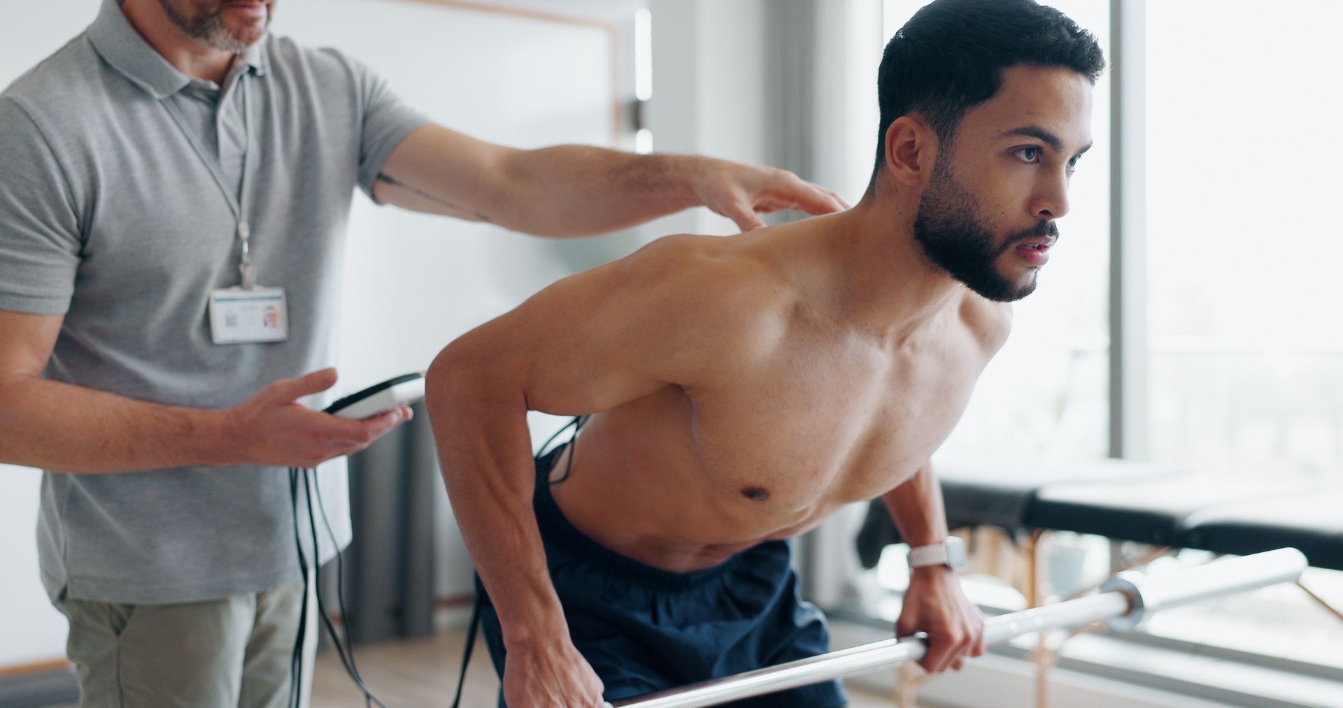 Man undergoing physical therapy with therapist. The man leans forward, holding a bar. A device is attached to his back.