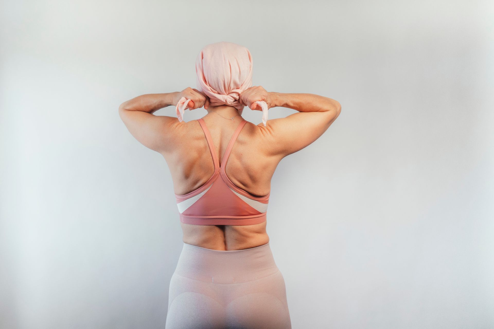 Person tying a pink scarf, wearing a pink bra and skirt, standing against a white wall.
