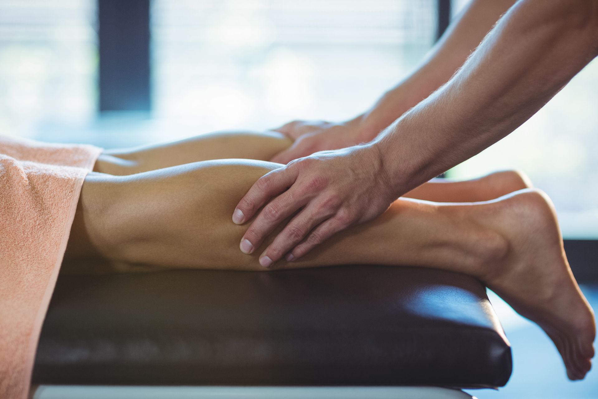 Hands massaging the lower legs of a person lying on a massage table, near a window
