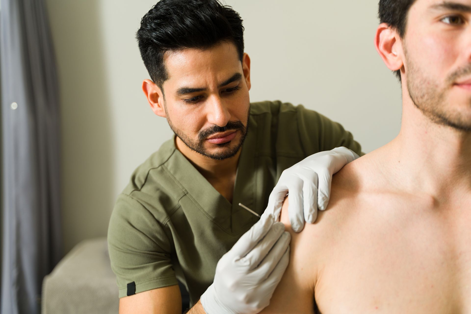 Doctor in gloves inserting a needle into a patient's shoulder. Neutral setting, focused expressions.
