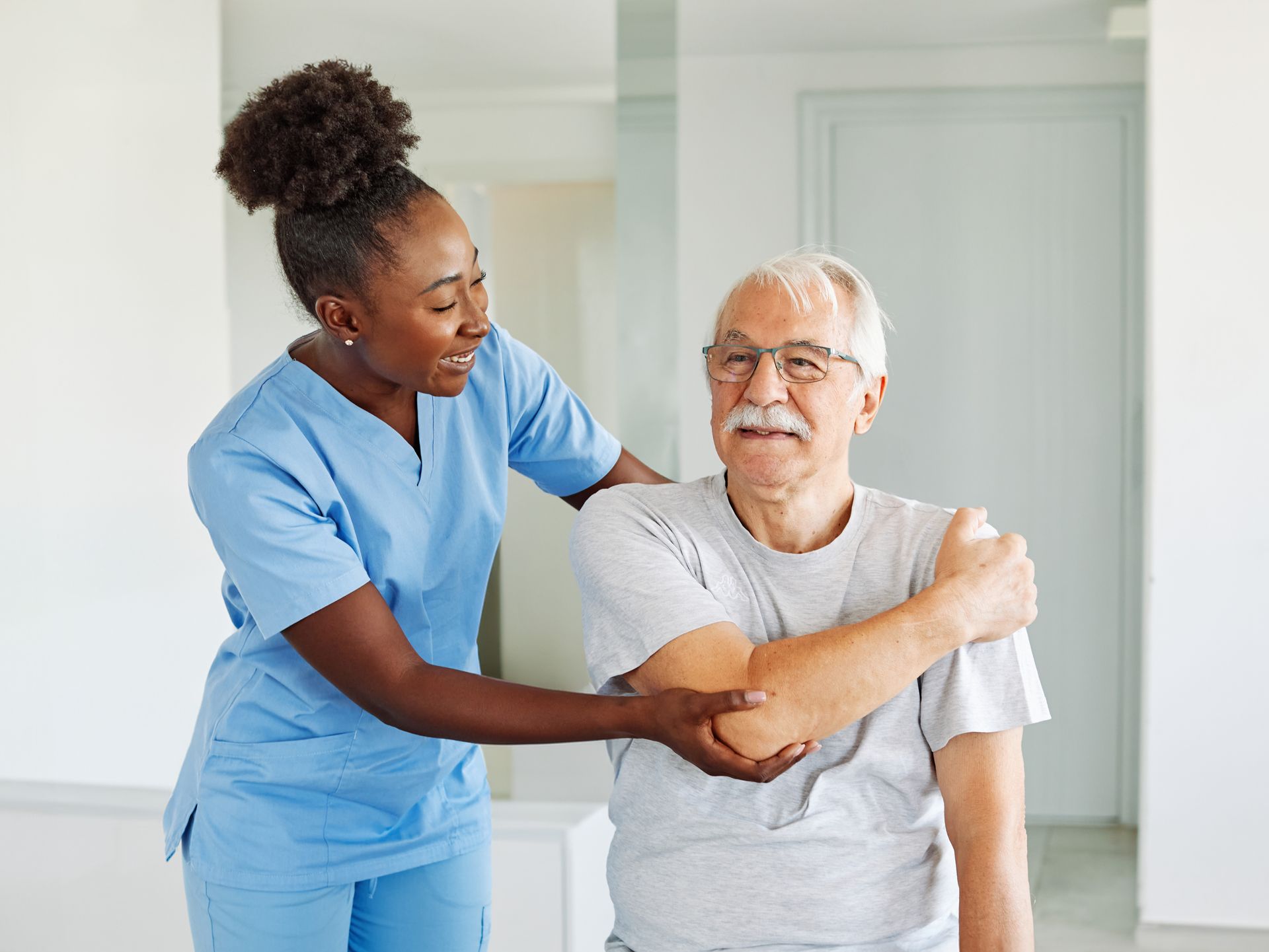 A healthcare worker in blue scrubs assists an older person in a gray shirt with arm movement, indoors.