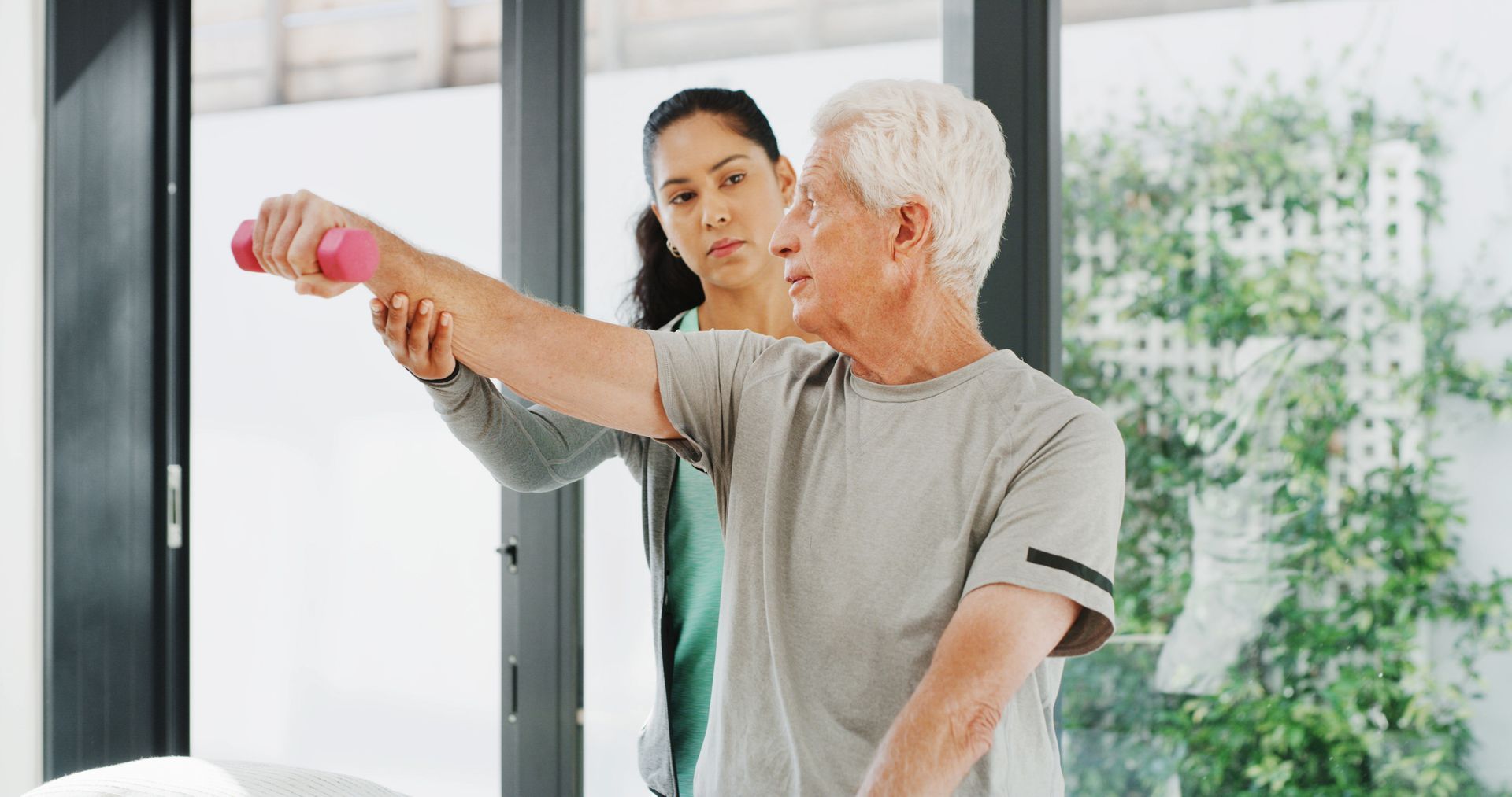 Therapist assists a person with gray hair lifting a pink dumbbell in a bright room.