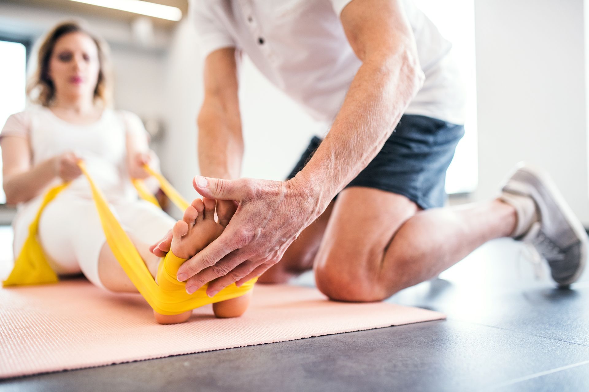 Therapist assisting a patient with foot exercises using a yellow resistance band on a pink mat.
