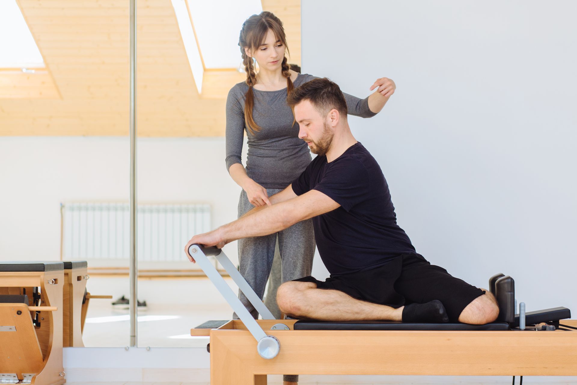 A professional instructor guides a person through a Pilates exercise on a reformer machine in a bright studio.