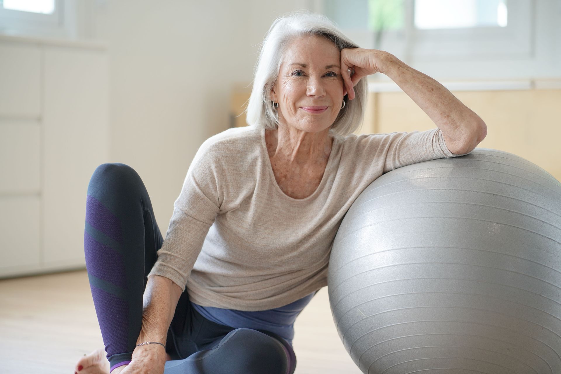 Woman resting by exercise ball, smiling. Indoors, neutral colors.