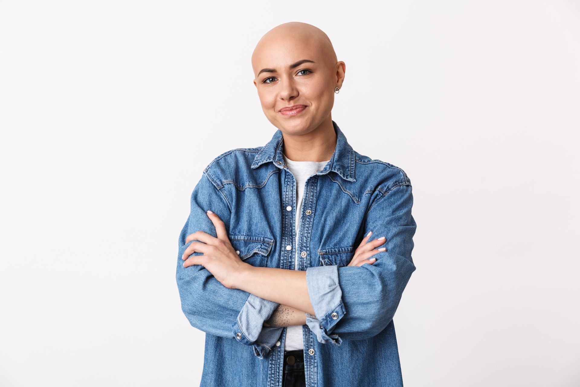 A smiling person with a shaved head, wearing a blue denim shirt, standing with arms crossed against a white background.