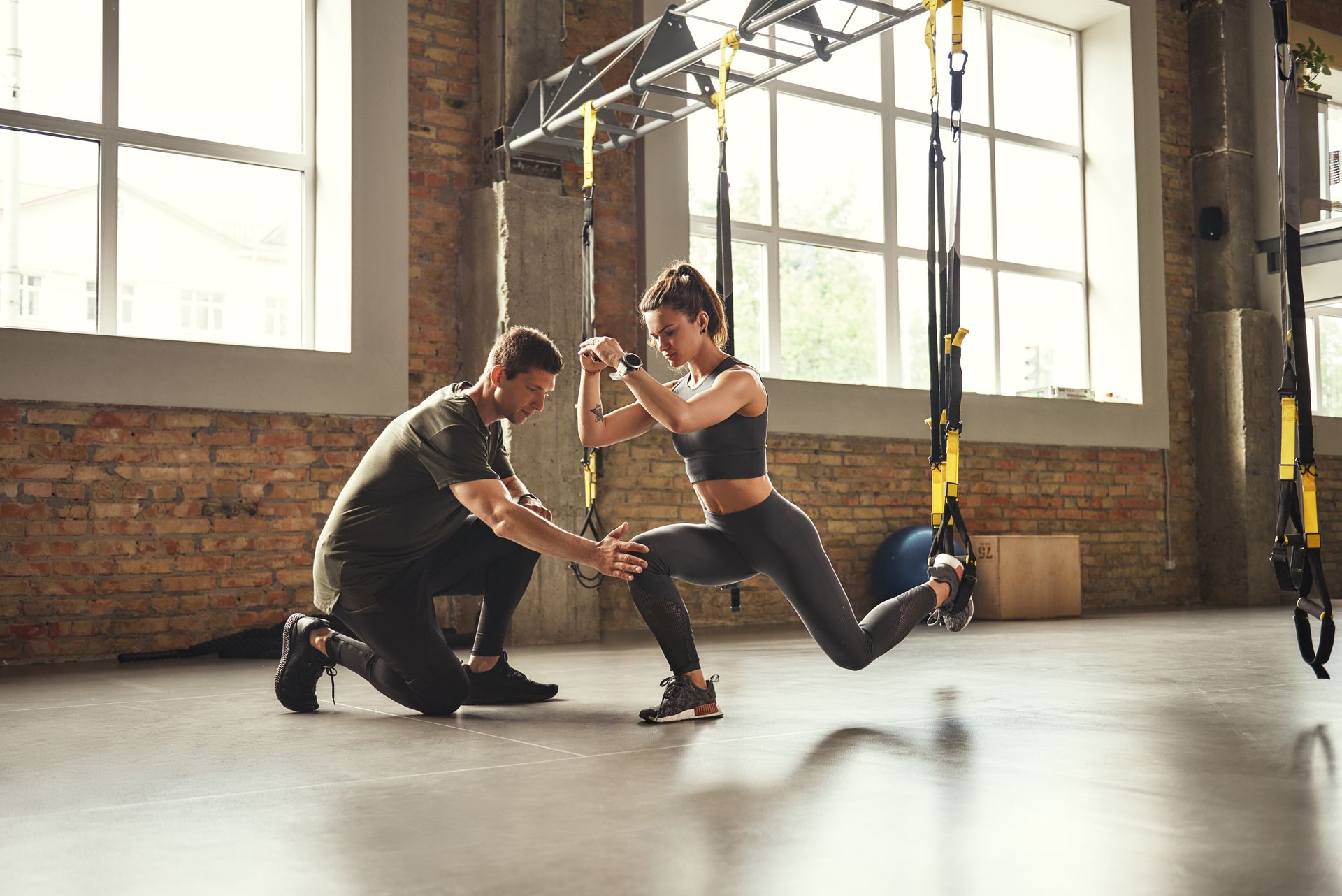 Woman doing a lunge with trainer. They are in a gym with TRX straps and natural light.