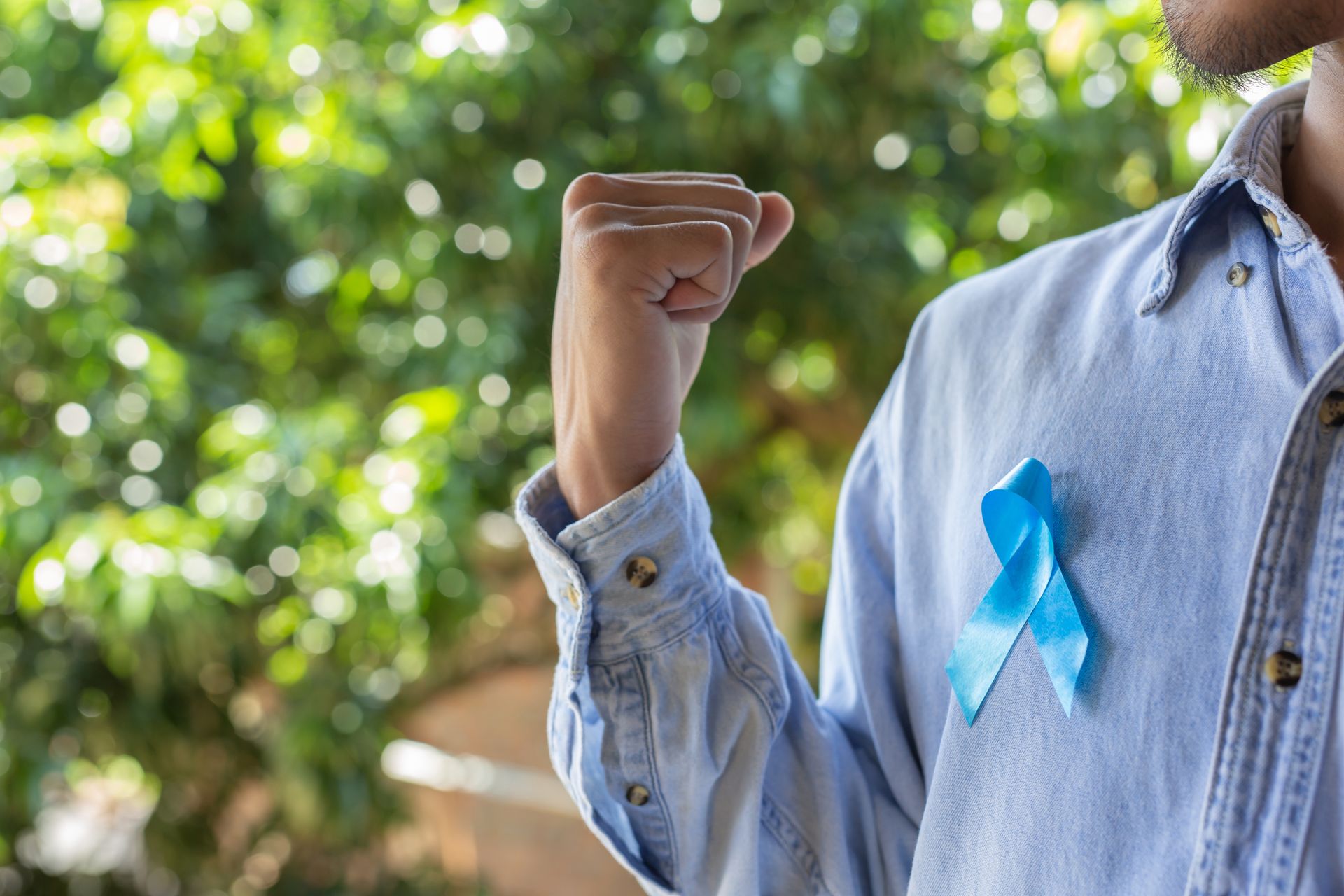 A person in a light blue denim shirt with a blue awareness ribbon pinned to their chest, flexing their arm.