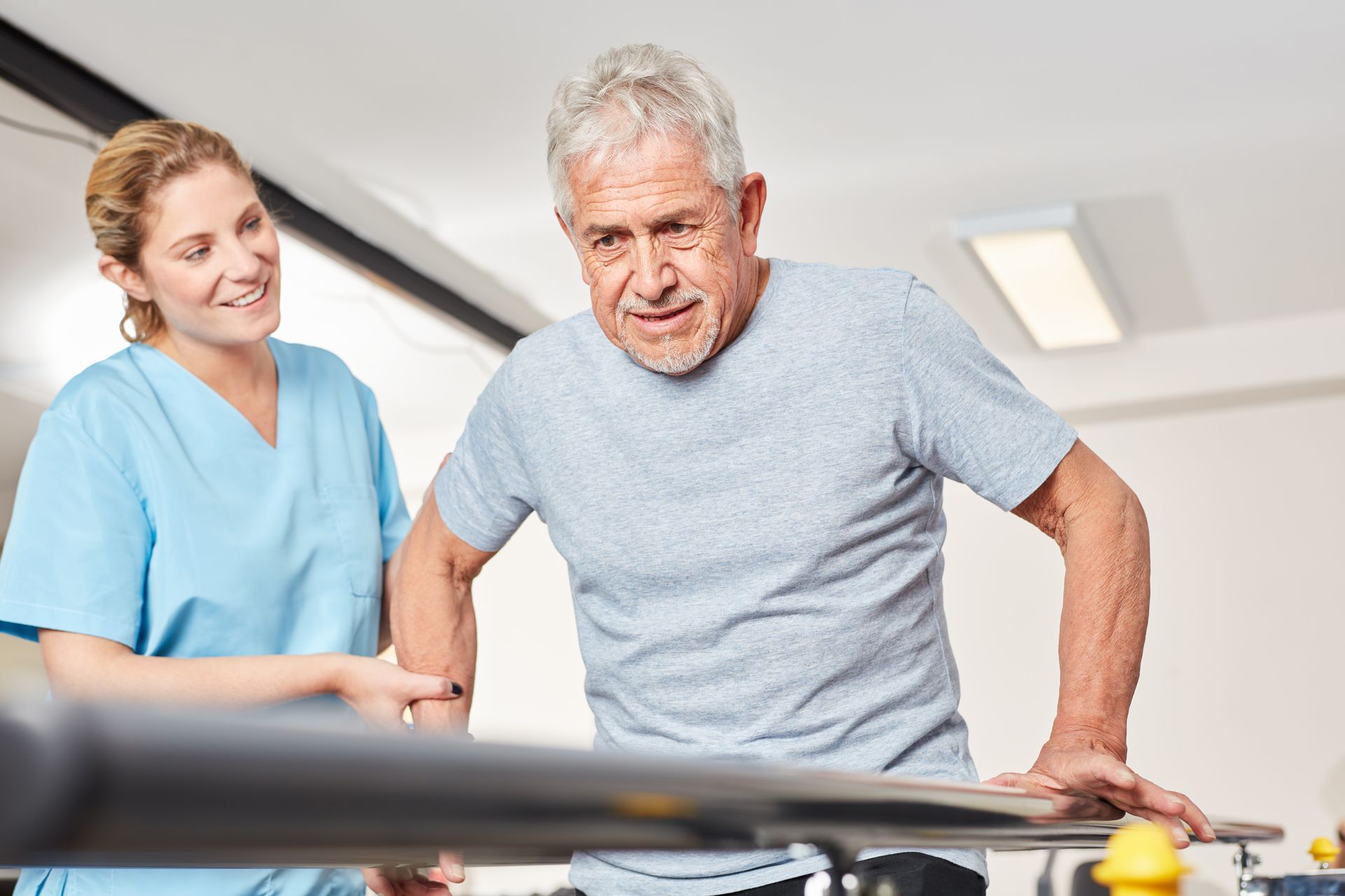 Woman in blue scrubs assists older man walking with a parallel bar in a physical therapy setting.