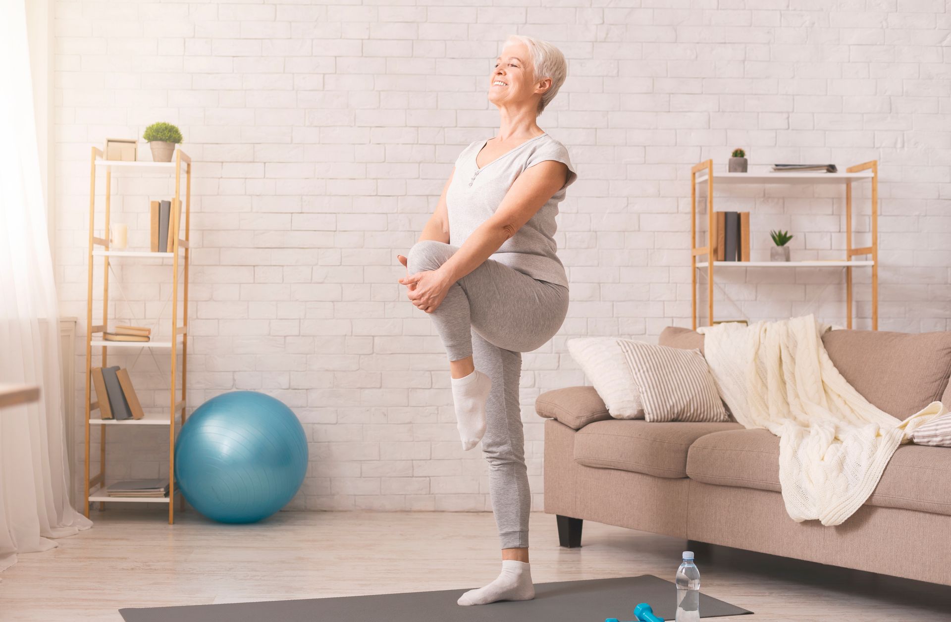 Woman stretching leg, holding knee, in light-filled living room. Blue exercise ball and sofa visible.