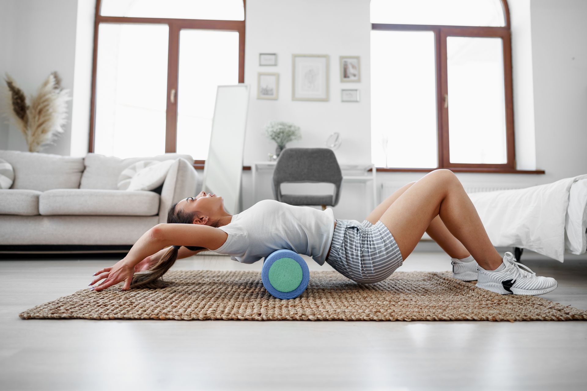 Woman doing back exercise on a foam roller, lying on a woven rug in a room.