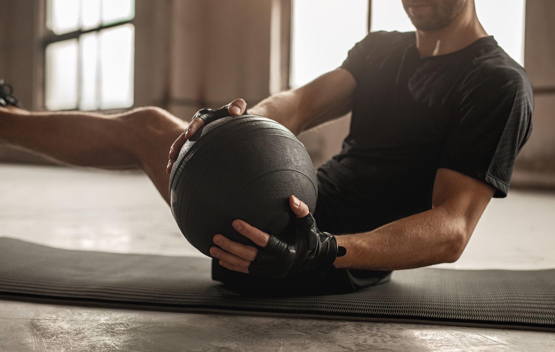 A person in a black t-shirt and workout gloves performs a seated medicine ball exercise on a gym mat.
