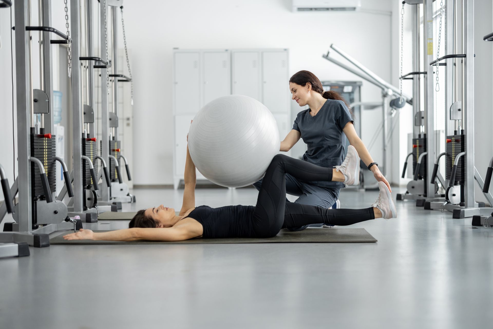 Woman on mat with ball, aided by another woman in a gym. Exercise.