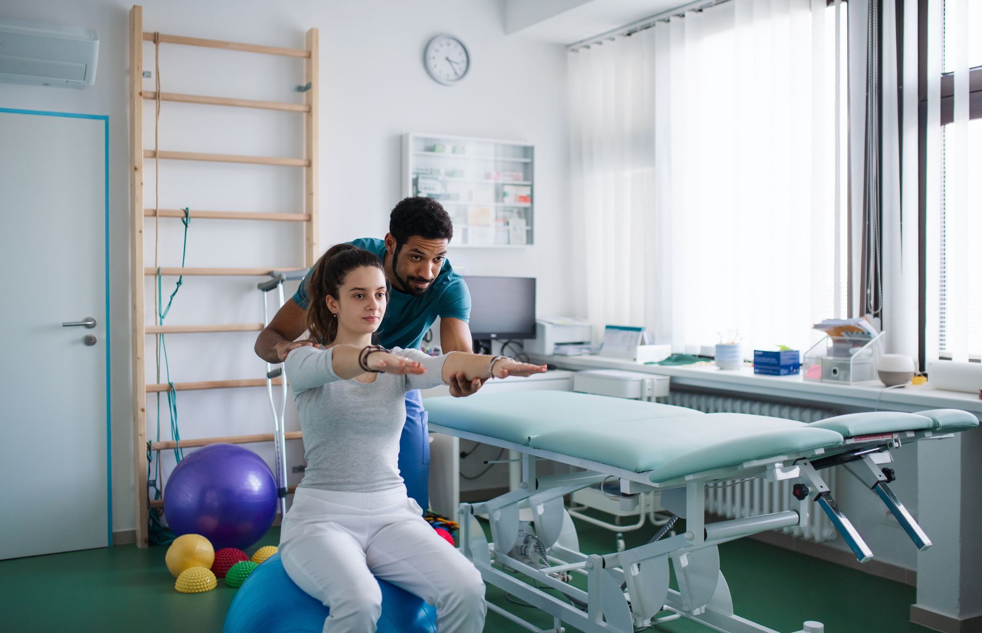 Therapist guiding a person sitting on a stability ball, arms outstretched, in a therapy room.