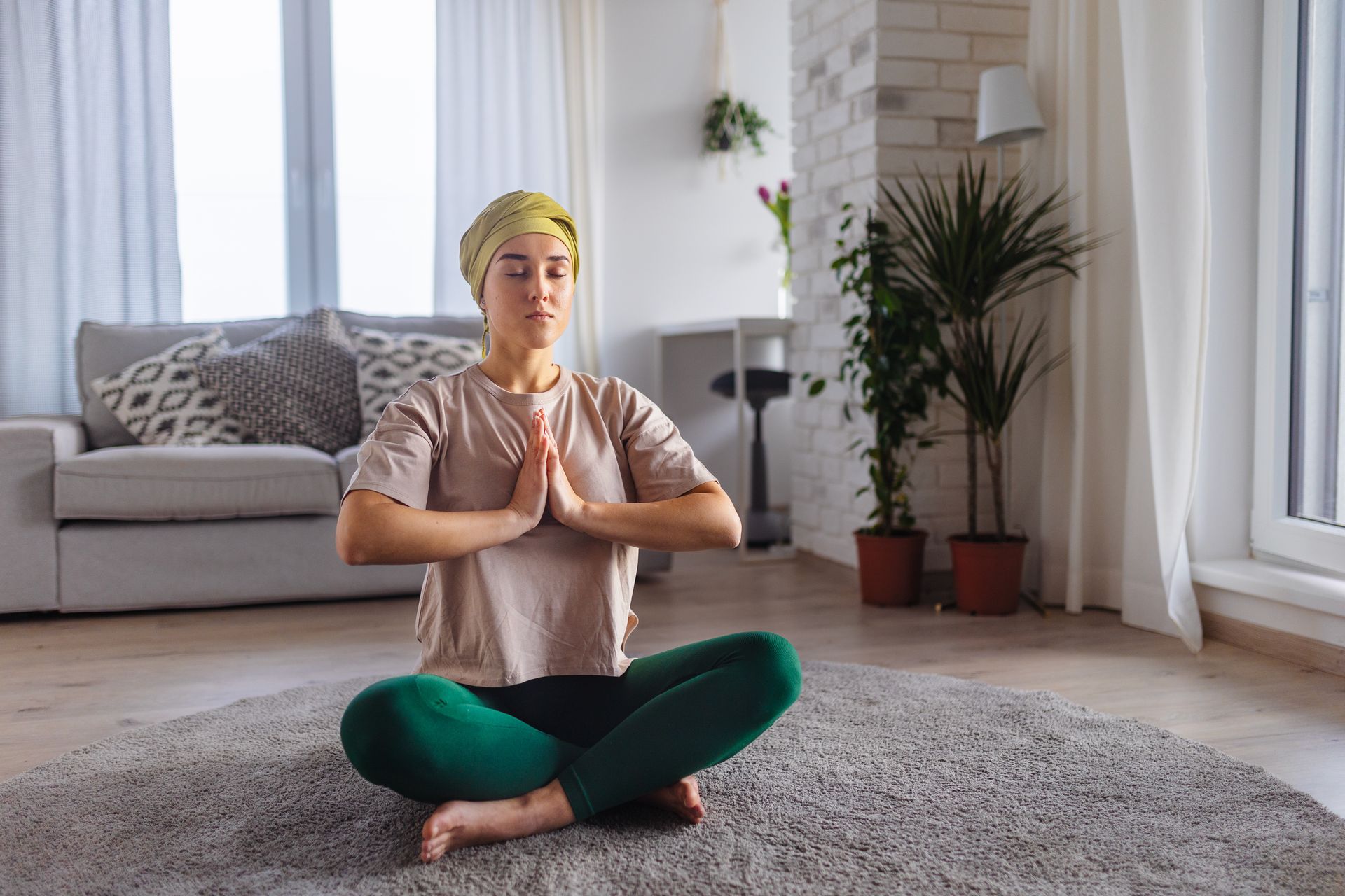 Woman in lotus pose, hands in prayer, eyes closed, on a rug in a living room, practicing yoga.