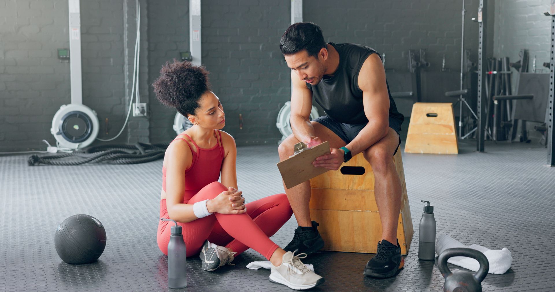 Woman and man in workout gear discussing at a gym; man holding clipboard, woman sitting with hand on knee.