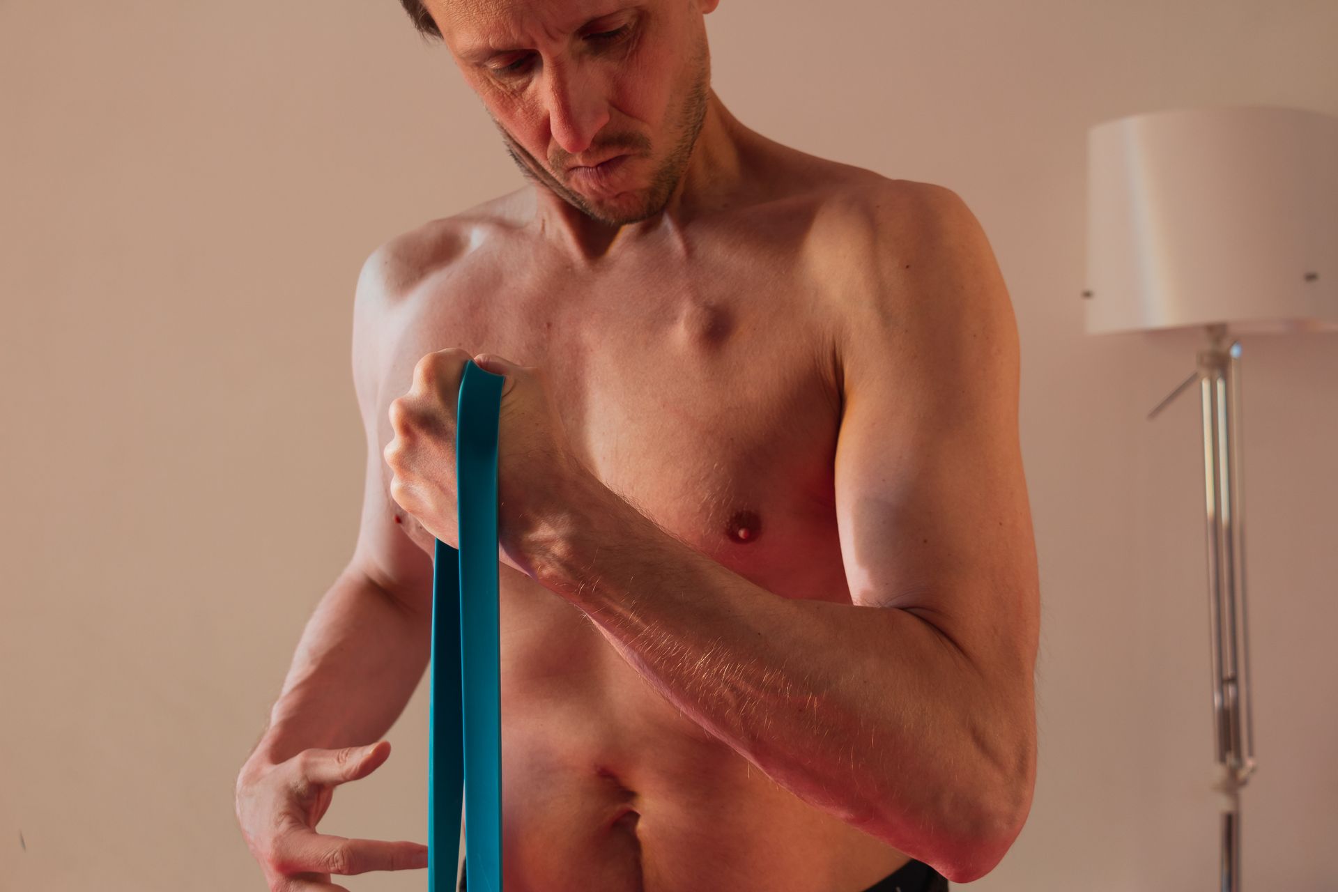 Man holding a blue resistance band, looking down. Setting is indoors, near a lamp.