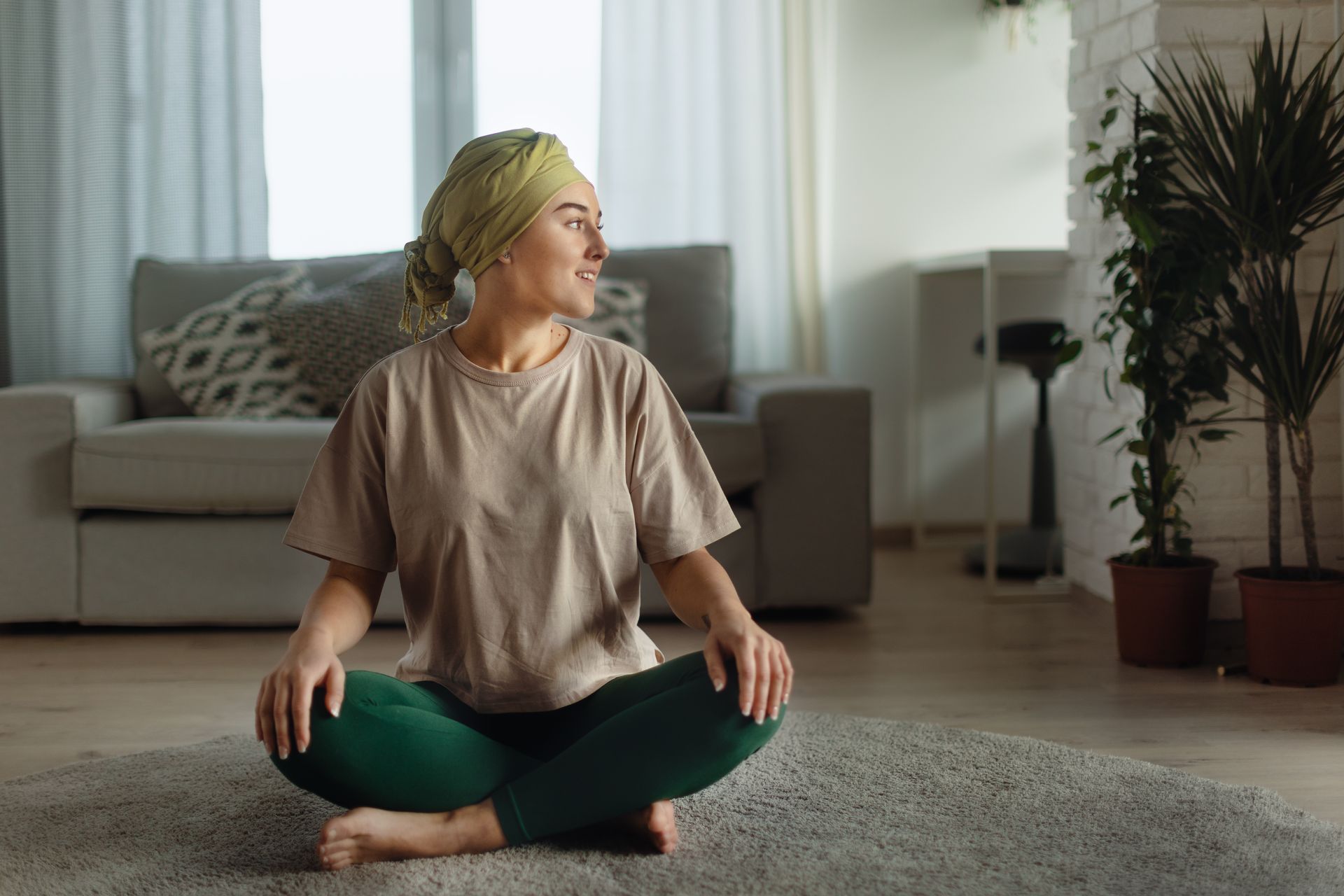 A person in a light green headwrap and beige t-shirt sits cross-legged on a circular rug, smiling while looking away.