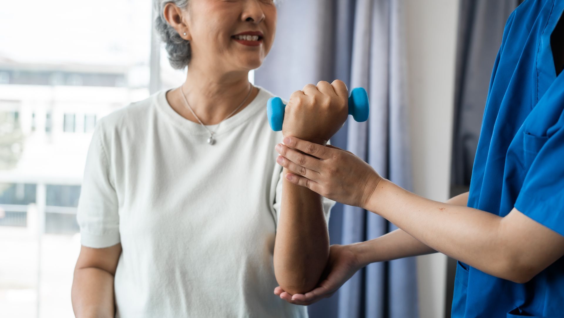 Woman doing arm exercises with a small dumbbell, assisted by a person in blue scrubs. Indoors, natural light.