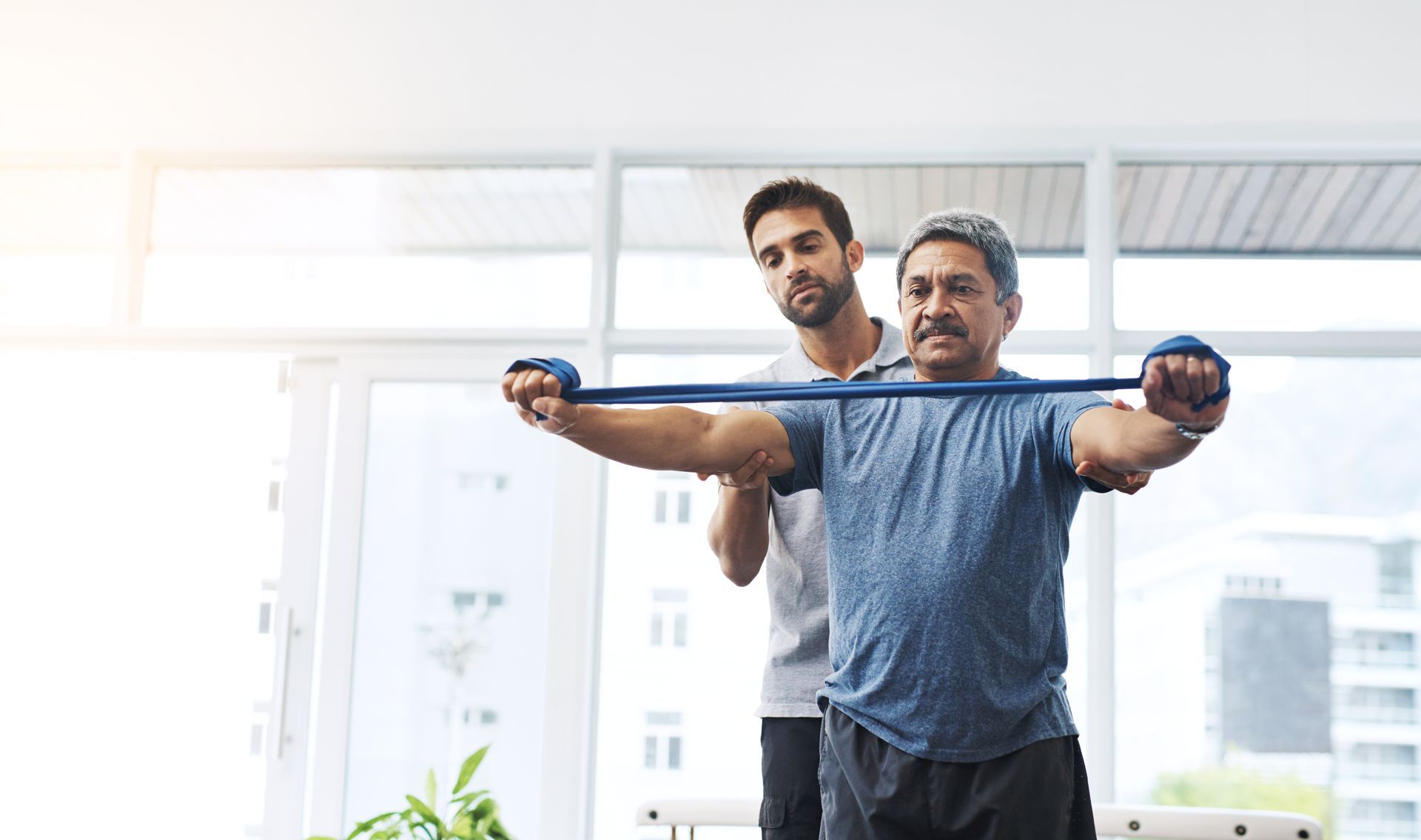 Therapist assisting a patient with arm exercises using a resistance band in a bright room.