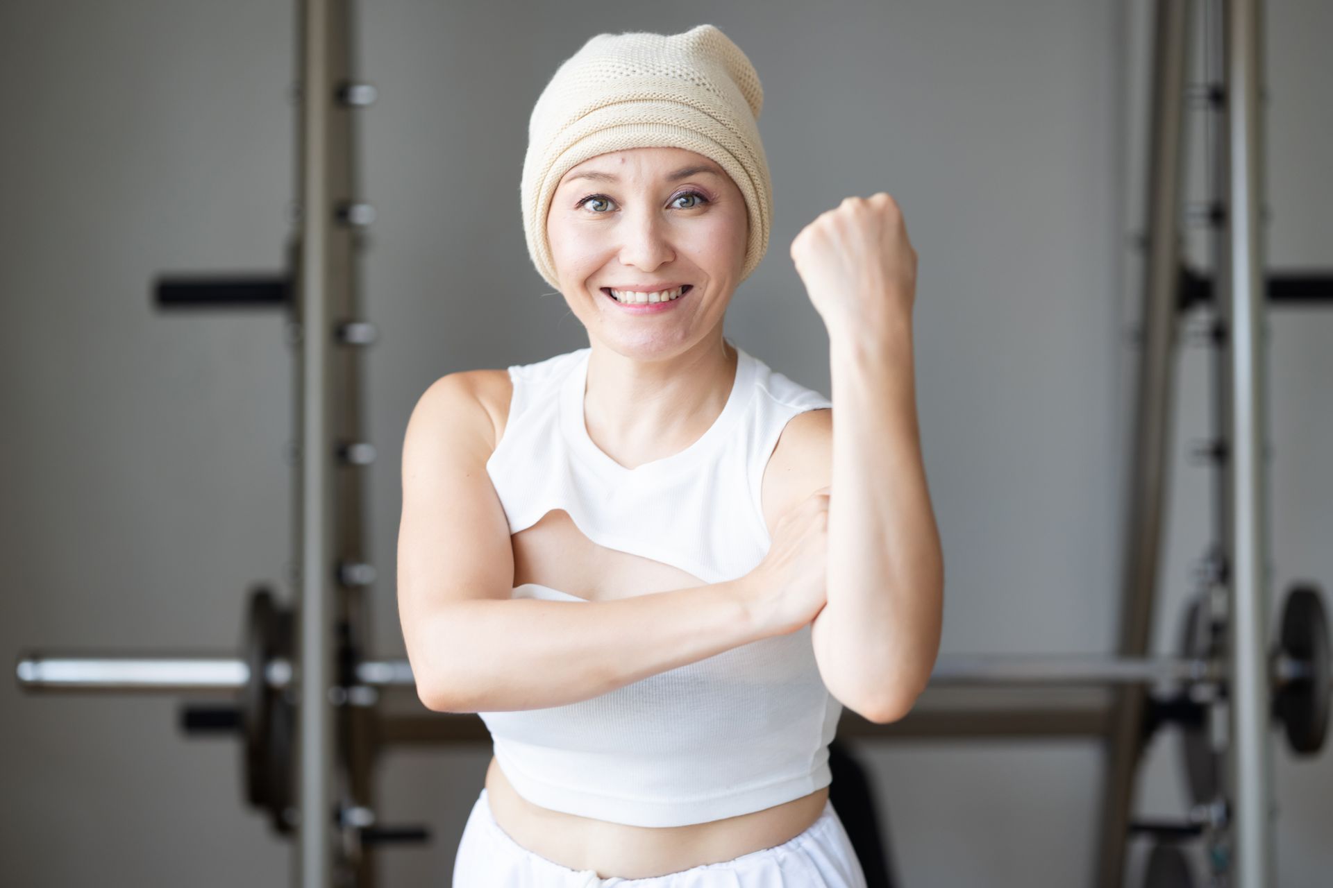 Woman in white outfit and beanie flexing arm in gym.