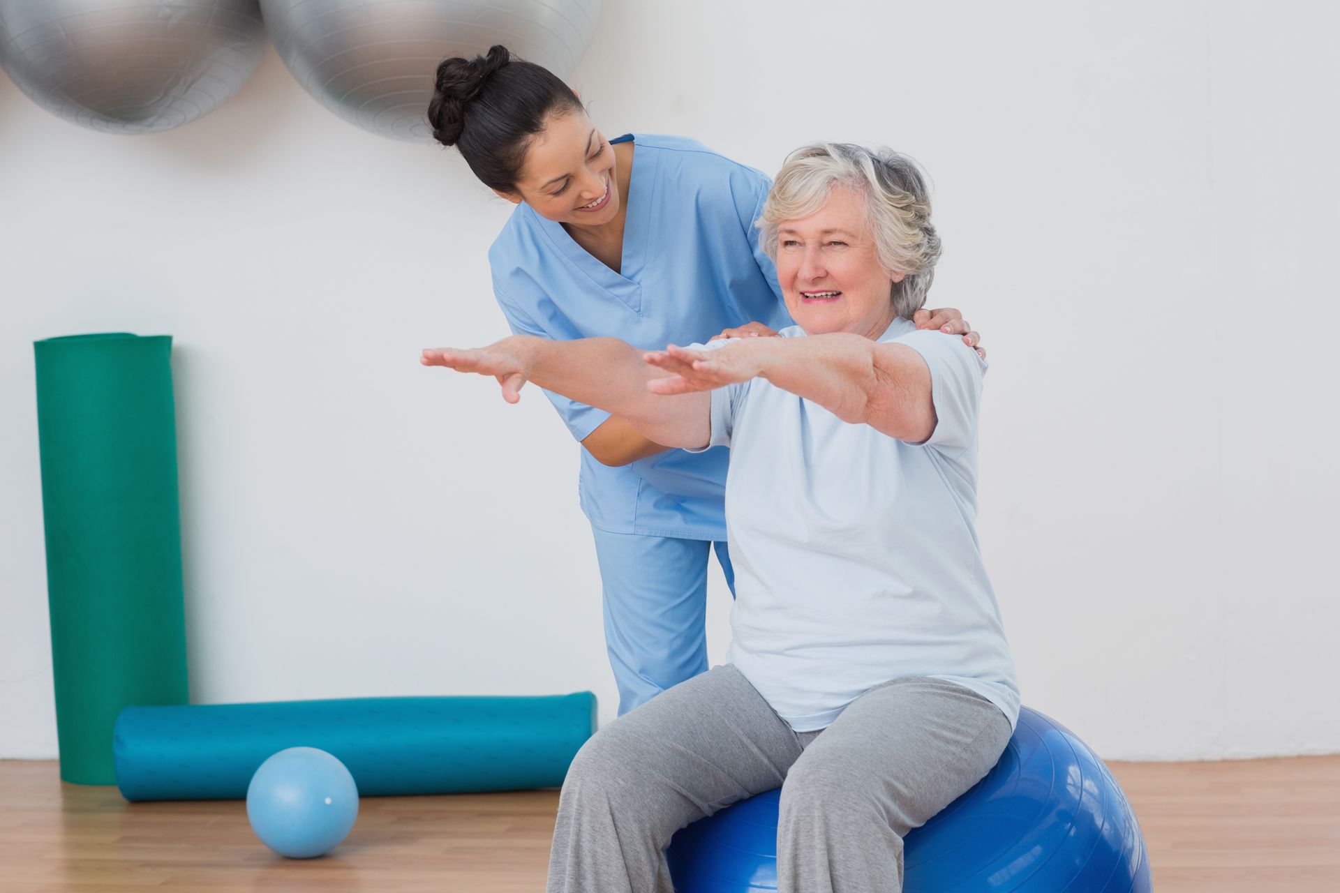 Therapist assists senior woman seated on exercise ball, arms outstretched, in a gym.