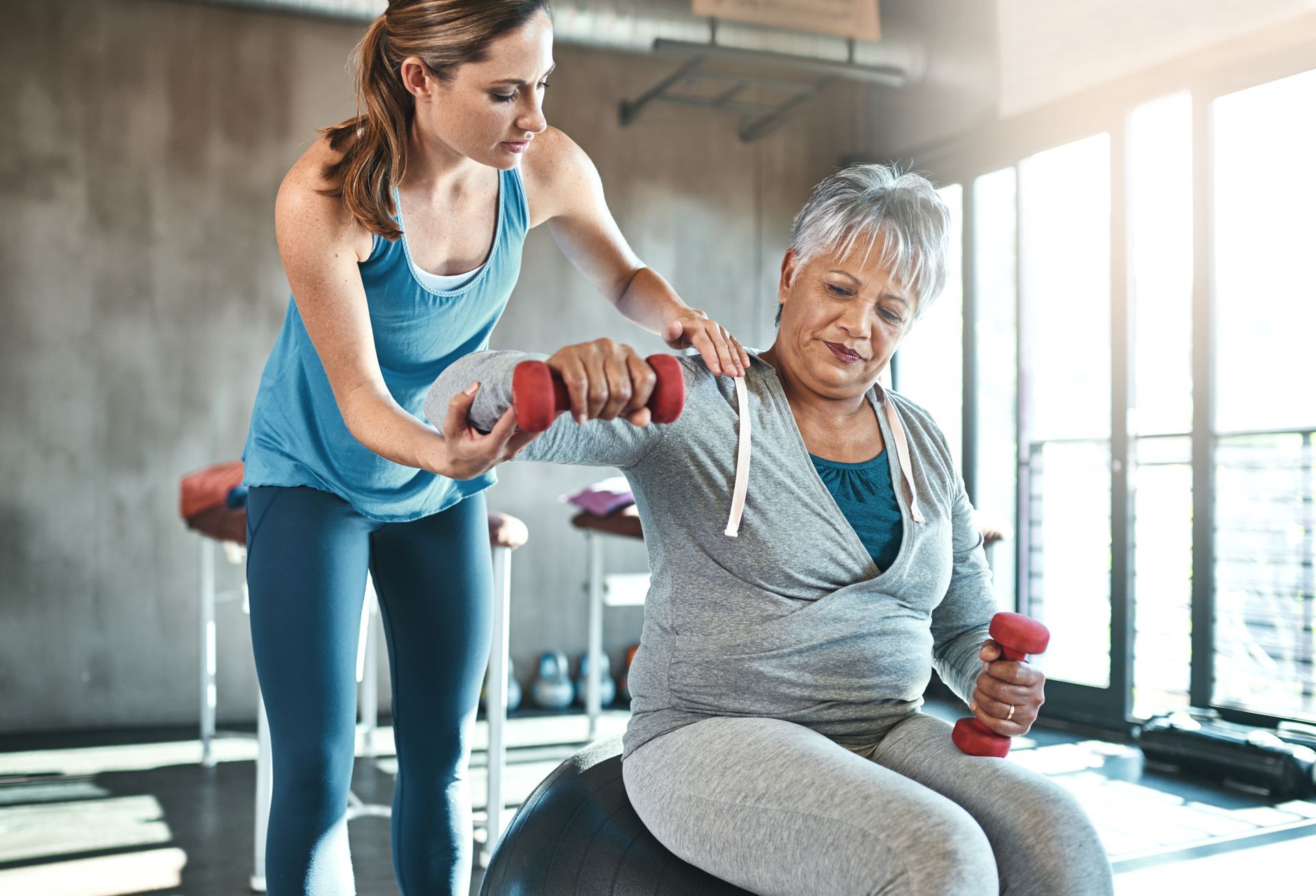 Trainer assisting a person doing dumbbell exercises while seated on a balance ball in a gym.