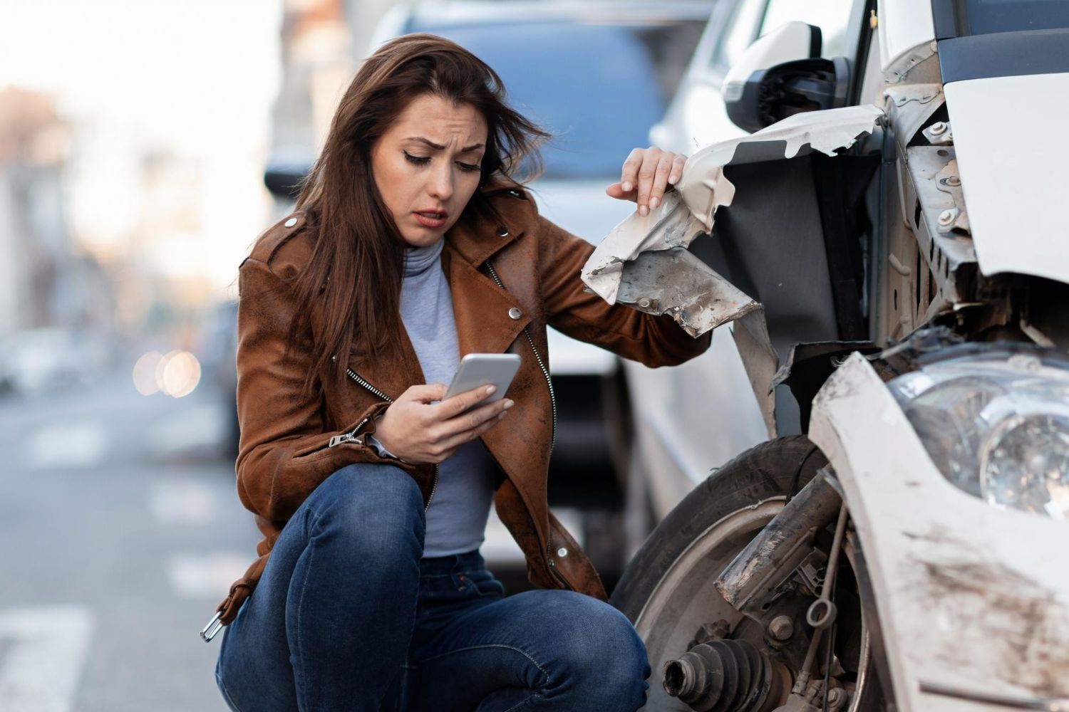 A woman is kneeling down next to a damaged car and looking at her phone.