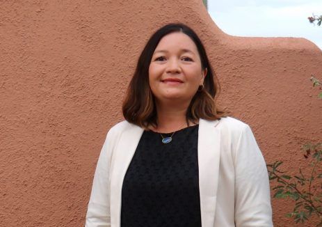 A woman in a white jacket and black top is standing in front of a red wall.