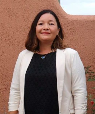 A woman in a black dress and white jacket is standing in front of a red wall.