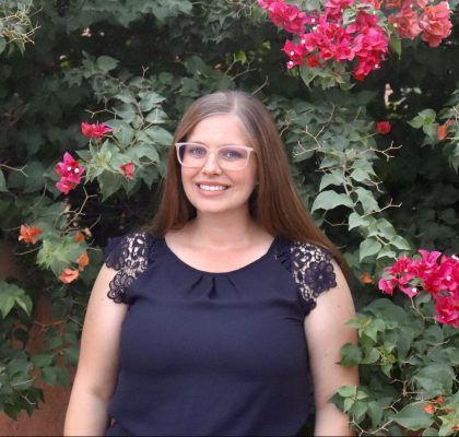 A woman wearing glasses is standing in front of a bush with pink flowers.