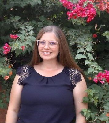 A woman wearing glasses is standing in front of a bush with pink flowers.