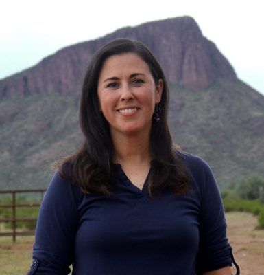 A woman in a blue shirt is smiling in front of a mountain.