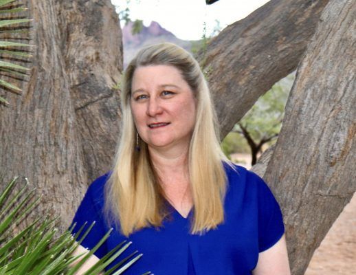 A woman in a blue shirt is standing in front of a tree.