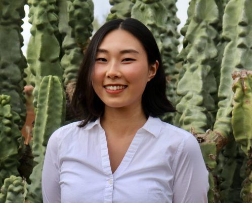 A woman in a white shirt is smiling in front of a cactus.