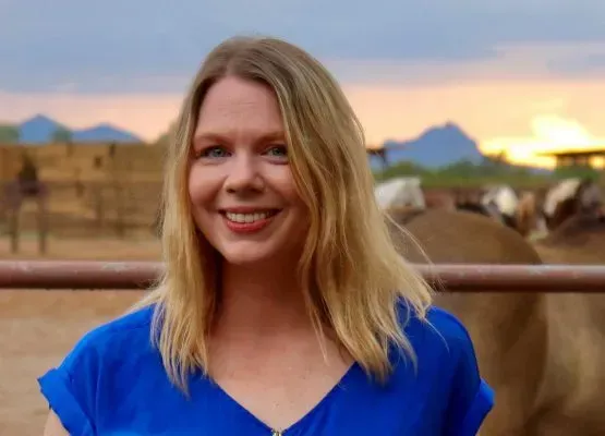 A woman in a blue shirt is smiling in front of a fence with cows in the background