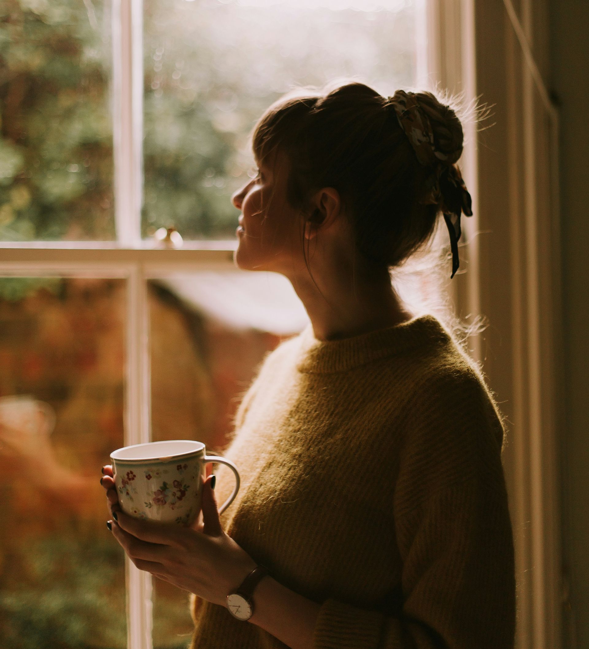 A woman in a yellow sweater is holding a cup of coffee in front of a window.